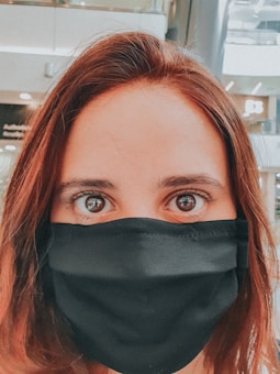 A close-up portrait of a person with long, brown hair, wearing a black face mask that covers their nose and mouth. The background appears to be an indoor setting, likely a mall or public space, indicated by blurred signage and bright lighting.