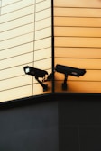 Two surveillance cameras are mounted on the corner of a building. The building features a modern design with a facade composed of horizontal panels in a soft yellow color. The cameras are positioned to cover different angles for optimal security coverage. The lower portion of the building is darker, providing a contrasting base.