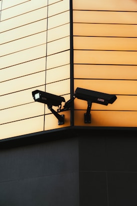 Two surveillance cameras are mounted on the corner of a building. The building features a modern design with a facade composed of horizontal panels in a soft yellow color. The cameras are positioned to cover different angles for optimal security coverage. The lower portion of the building is darker, providing a contrasting base.