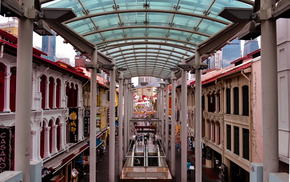 A bustling street scene with a glass canopy overhead, flanked by colorful traditional buildings with ornate fa&ccedil;ades. People walk along the street, and signs with Chinese characters are displayed prominently. Modern skyscrapers can be glimpsed in the background, contrasting with the historical architecture.