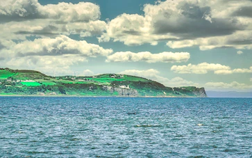 A scenic view of a coastline with vibrant green fields stretching across the landscape under a sky filled with fluffy clouds. The turquoise sea in the foreground contrasts with the greenery of the land. In the background, wind turbines can be seen on the hills.