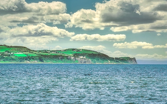 A scenic view of a coastline with vibrant green fields stretching across the landscape under a sky filled with fluffy clouds. The turquoise sea in the foreground contrasts with the greenery of the land. In the background, wind turbines can be seen on the hills.