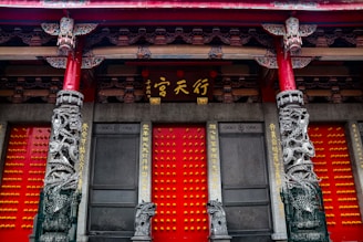 An ornate architectural facade with vibrant red doors featuring rows of golden studs. Intricately carved stone columns with dragon motifs flank the entrance. Above the doors, there's detailed woodwork with traditional Chinese characters written on panels.