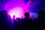 Silhouettes of people dance in a dimly lit environment filled with vibrant purple and blue lights, creating a lively and energetic atmosphere. Some of the individuals have their hands raised, as if enjoying the music and ambiance.