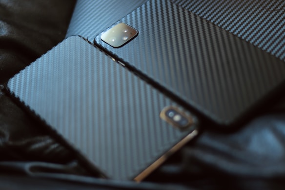 Two electronic devices featuring carbon fiber textured back covers laid on a dark surface. The devices appear to be smartphones or tablets, with camera modules visible on the top left corner.