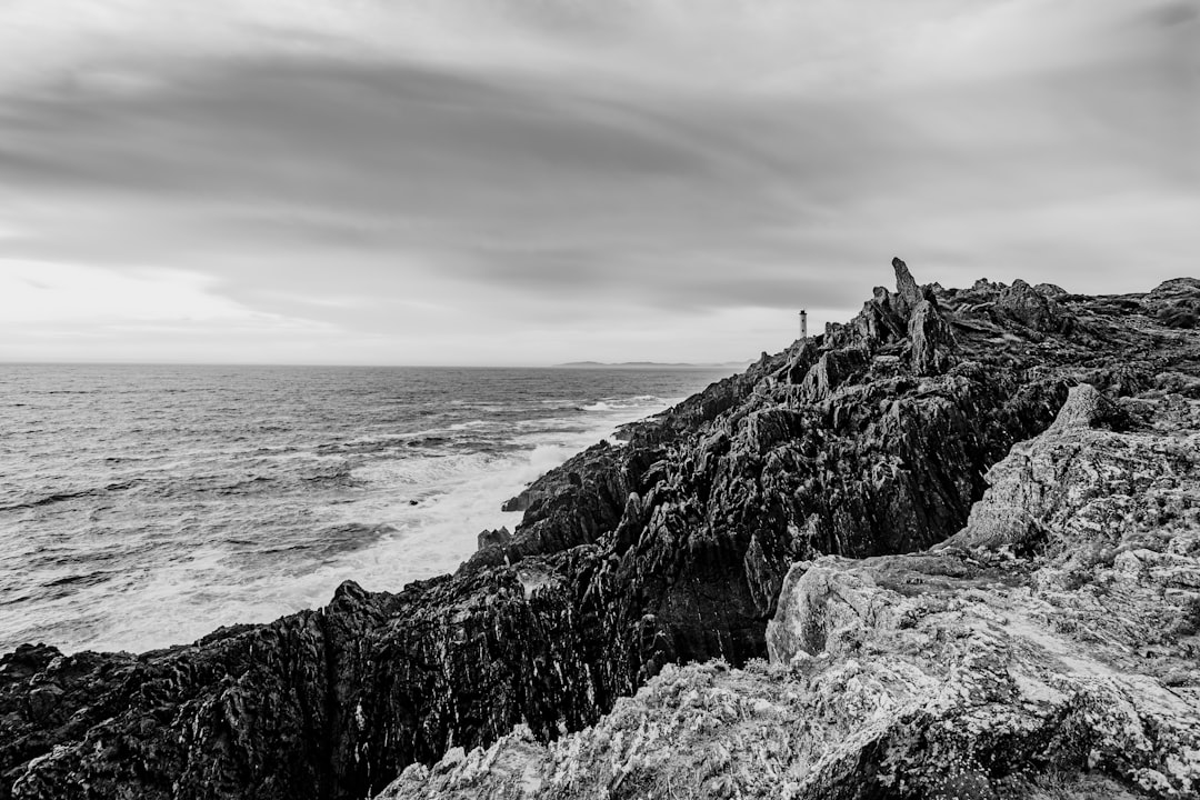 grayscale photo of person standing on rock formation near sea
