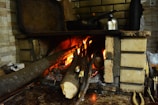 A rustic indoor fireplace made of bricks, with several logs burning inside. The fire is bright and produces a warm glow, illuminating the surrounding area. On top of the fireplace, there are a kettle and a few other items, suggesting a cozy and homely environment.