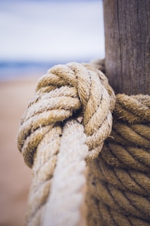 A close-up view of a thick, coarse rope tightly wrapped and knotted around a wooden post. The texture of the rope is rough, with visible fibers, and the wood appears weathered. The background is blurred, suggesting an outdoor setting.
