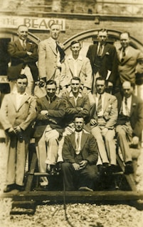 A group of men dressed in suits and ties are arranged in two rows, some standing and others sitting on a wooden structure. The background shows part of an old brick building with an arched window and a sign that reads 'BEACH'. The image has a sepia tone, indicating it might be vintage.
