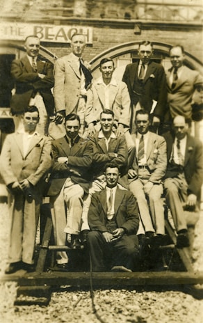 A group of men dressed in suits and ties are arranged in two rows, some standing and others sitting on a wooden structure. The background shows part of an old brick building with an arched window and a sign that reads 'BEACH'. The image has a sepia tone, indicating it might be vintage.