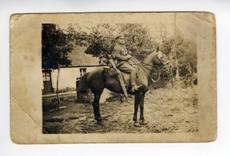 A vintage sepia-toned photograph featuring a person in military attire seated on a horse. The background includes a rustic building with a tiled roof partially obscured by trees. The ground appears rough and natural, suggesting a rural setting.