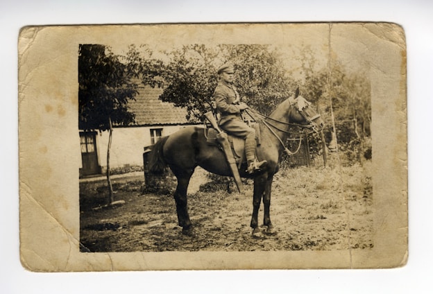 A vintage sepia-toned photograph featuring a person in military attire seated on a horse. The background includes a rustic building with a tiled roof partially obscured by trees. The ground appears rough and natural, suggesting a rural setting.