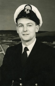 A man in a naval uniform with a double-breasted jacket and a peaked cap, featuring an emblem on the hat. He is posing for a portrait with a neutral expression, and the background shows an abstract or possibly maritime scene.