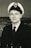 A man in a naval uniform with a double-breasted jacket and a peaked cap, featuring an emblem on the hat. He is posing for a portrait with a neutral expression, and the background shows an abstract or possibly maritime scene.