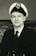 A man in a naval uniform with a double-breasted jacket and a peaked cap, featuring an emblem on the hat. He is posing for a portrait with a neutral expression, and the background shows an abstract or possibly maritime scene.