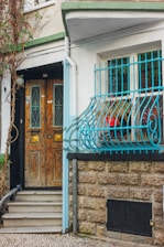 A rustic wooden door with decorative metalwork sits beside a window adorned with blue iron bars. The stone wall is textured, contrasting with the brick pathway leading to the step. Vines climb the side of the building, adding a touch of nature. Red flowers can be seen in a planter behind the window grill.
