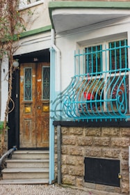 A rustic wooden door with decorative metalwork sits beside a window adorned with blue iron bars. The stone wall is textured, contrasting with the brick pathway leading to the step. Vines climb the side of the building, adding a touch of nature. Red flowers can be seen in a planter behind the window grill.