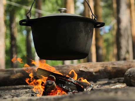 A close-up of sizzling campfire stew bubbling in a rustic cast iron pot surrounded by forest greenery.