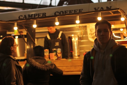 A cozy coffee van is illuminated by several warm light bulbs. A barista, wearing a dark apron, is behind the counter. Three people stand outside, one closer to the camera with a hoodie, while the other two are at the counter possibly ordering.