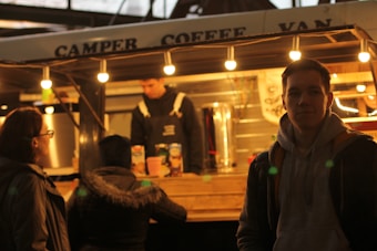 A cozy coffee van is illuminated by several warm light bulbs. A barista, wearing a dark apron, is behind the counter. Three people stand outside, one closer to the camera with a hoodie, while the other two are at the counter possibly ordering.