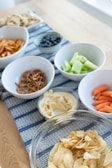 An assortment of snacks is arranged on a wooden table with a blue patterned runner. There are bowls containing potato chips, pretzels, hummus with a side of celery, blueberries, baby carrots, and a tray of cookies in the background.
