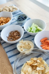 An assortment of snacks is arranged on a wooden table with a blue patterned runner. There are bowls containing potato chips, pretzels, hummus with a side of celery, blueberries, baby carrots, and a tray of cookies in the background.
