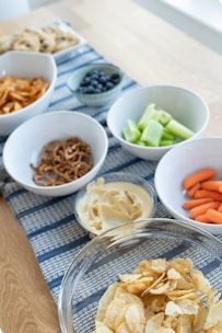 An assortment of snacks is arranged on a wooden table with a blue patterned runner. There are bowls containing potato chips, pretzels, hummus with a side of celery, blueberries, baby carrots, and a tray of cookies in the background.