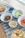 An assortment of snacks is arranged on a wooden table with a blue patterned runner. There are bowls containing potato chips, pretzels, hummus with a side of celery, blueberries, baby carrots, and a tray of cookies in the background.