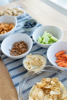 An assortment of snacks is arranged on a wooden table with a blue patterned runner. There are bowls containing potato chips, pretzels, hummus with a side of celery, blueberries, baby carrots, and a tray of cookies in the background.