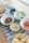 An assortment of snacks is arranged on a wooden table with a blue patterned runner. There are bowls containing potato chips, pretzels, hummus with a side of celery, blueberries, baby carrots, and a tray of cookies in the background.