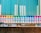 A row of colorful spray paint cans is lined up neatly against a backdrop of blue vertical blinds. The cans display a variety of colors including orange, yellow, green, blue, and pink. They are evenly spaced on a metallic shelf that contrasts with the textured brick wall below.