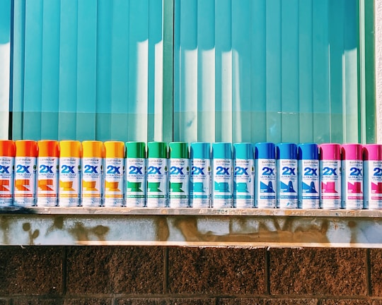 A row of colorful spray paint cans is lined up neatly against a backdrop of blue vertical blinds. The cans display a variety of colors including orange, yellow, green, blue, and pink. They are evenly spaced on a metallic shelf that contrasts with the textured brick wall below.