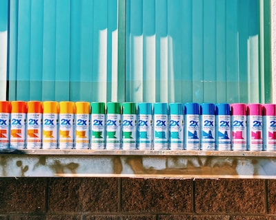 A row of colorful spray paint cans is lined up neatly against a backdrop of blue vertical blinds. The cans display a variety of colors including orange, yellow, green, blue, and pink. They are evenly spaced on a metallic shelf that contrasts with the textured brick wall below.