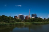 A cityscape with modern high-rise buildings set against a bright blue sky. A construction crane is visible, indicating ongoing development. Lush greenery and a calm water body are in the foreground, contrasting with the urban structures.