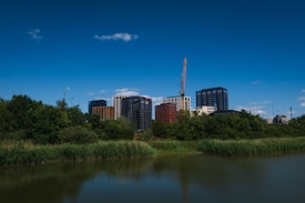 A cityscape with modern high-rise buildings set against a bright blue sky. A construction crane is visible, indicating ongoing development. Lush greenery and a calm water body are in the foreground, contrasting with the urban structures.