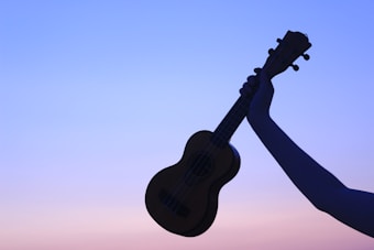 A silhouetted hand holds up a ukulele against the backdrop of a gradient sky transitioning from blue to pink.