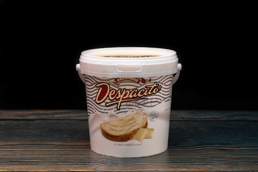 A white plastic container with a handle, labeled 'Despacito White Chocolate Spread,' featuring an image of a slice of toast topped with white chocolate spread. The container rests on a wooden surface against a dark background, highlighting its design.