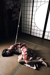 A person wearing a traditional red and white patterned kimono lies on a tatami mat floor. The room includes a shoji screen with a large circular window, creating a contrast of light and shadow. A rope is secured around the person's torso, adding a dynamic tension to the scene.