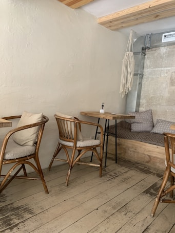 A cozy corner of a cafe features wooden chairs with cushions arranged around small tables. The floor is made of rustic wooden planks, and there is a seating area with a hanging macram&eacute; ornament on the wall. Neutral tones dominate the decor, with a focus on simple, natural materials.