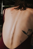 A close-up view of a person's back with visible spine details and a floral tattoo on the lower right side. The person has dark hair falling over their neck and is wearing a dark maroon garment.
