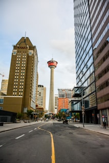 A city street stretches toward a tall observation tower in the distance, surrounded by modern high-rise buildings. The tower stands prominently with a red and white top section. On the left, a building with the HYATT logo is visible. The road is lined with trees and a few people are visible on the sidewalks.