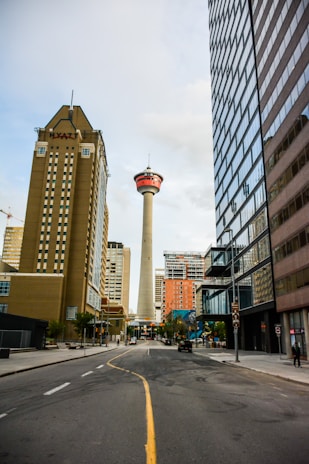 A city street stretches toward a tall observation tower in the distance, surrounded by modern high-rise buildings. The tower stands prominently with a red and white top section. On the left, a building with the HYATT logo is visible. The road is lined with trees and a few people are visible on the sidewalks.
