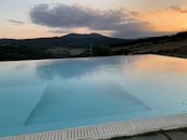 An infinity pool with a serene, glass-like surface reflecting the colorful hues of a sunset. Beyond the pool, rolling hills and a wooded landscape stretch toward the horizon. The sky displays vibrant shades of orange and pink through scattered clouds.