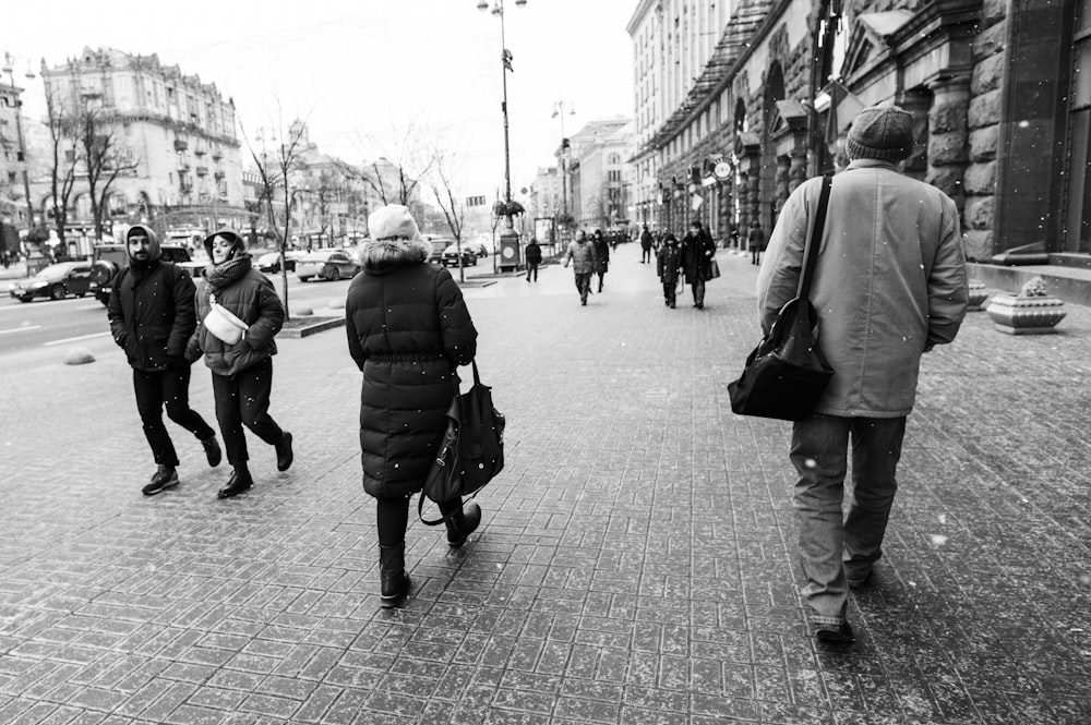grayscale photo of man in black jacket carrying backpack walking on sidewalk