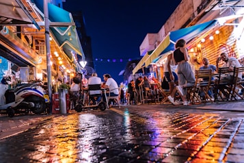 A lively street scene at night with numerous people sitting at outdoor tables of a caf&eacute; or restaurant. Vibrant lights from the venue and colorful reflections on the wet cobblestone street add a dynamic and energetic atmosphere. The awnings and surrounding buildings frame the buzzing urban environment.