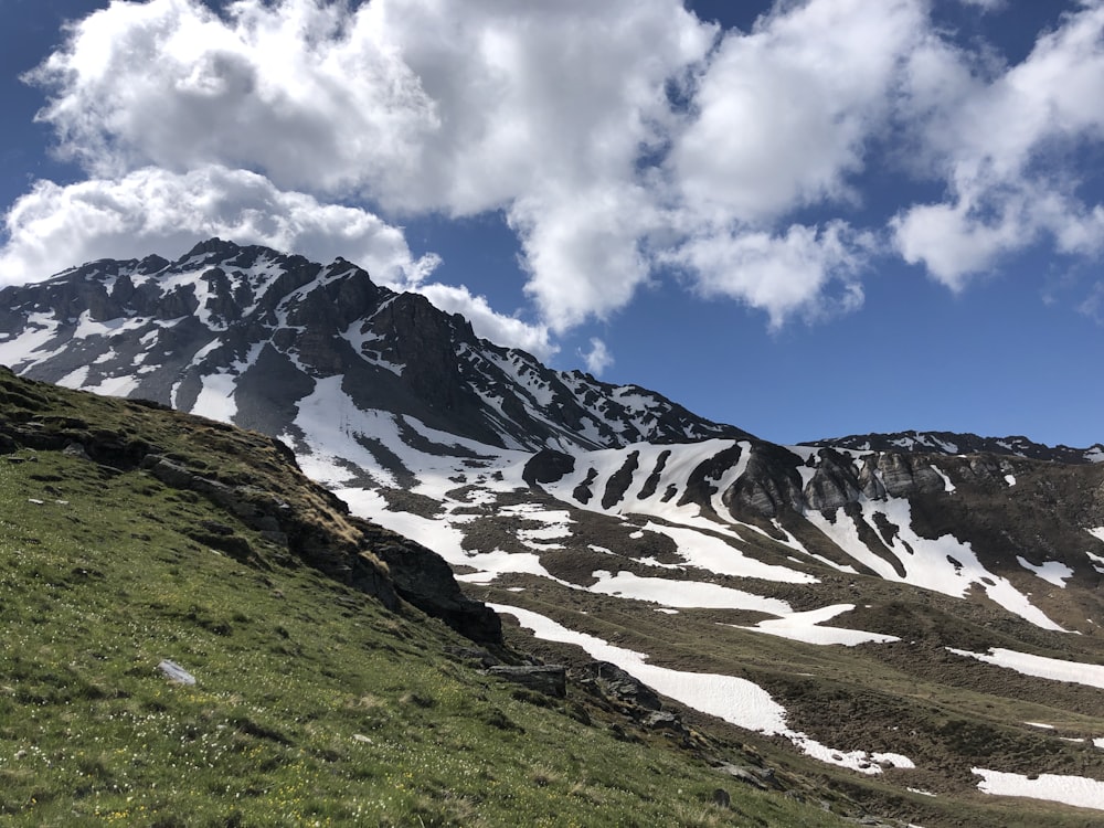 campo de grama verde perto da montanha coberta de neve sob nuvens brancas e céu azul durante o dia