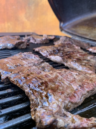 Juicy cuts of meat are being grilled on a barbecue grill. The meat appears to be well-seasoned and cooked, with a slight sheen indicating it is still being grilled. In the background, there is an open grill lid and a blurred out backdrop with warm colors.