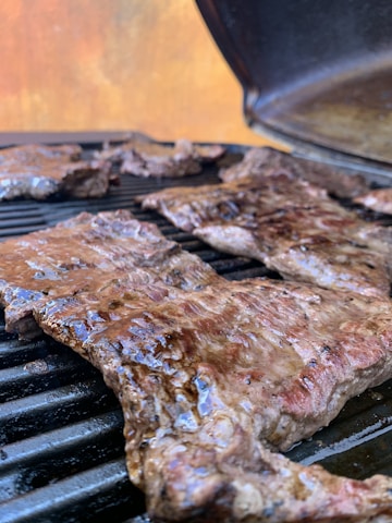 Juicy cuts of meat are being grilled on a barbecue grill. The meat appears to be well-seasoned and cooked, with a slight sheen indicating it is still being grilled. In the background, there is an open grill lid and a blurred out backdrop with warm colors.