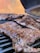 Juicy cuts of meat are being grilled on a barbecue grill. The meat appears to be well-seasoned and cooked, with a slight sheen indicating it is still being grilled. In the background, there is an open grill lid and a blurred out backdrop with warm colors.