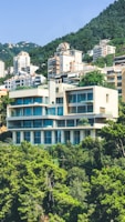 A modern, multi-story building nestled amongst lush green trees and set against a mountainous backdrop. The building features large windows and balconies, with a minimalist architectural design. Surrounding the main building are other residential structures set on the hillside.